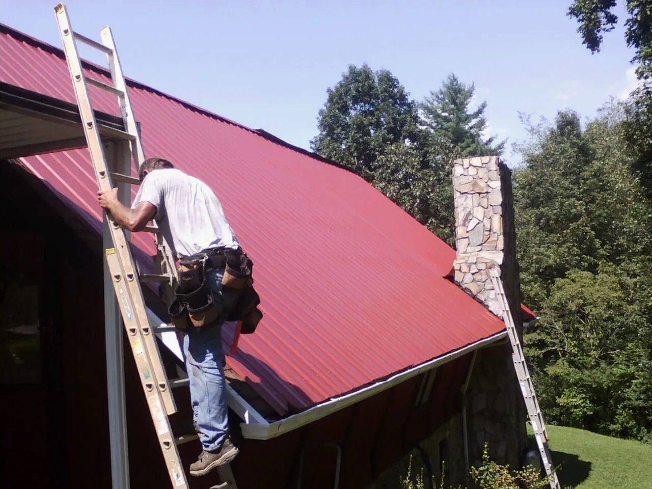 Roofer on a ladder beside a red metal roof and stone chimney — Soffit & Fascia Repair by Roofing Brothers in Aiken